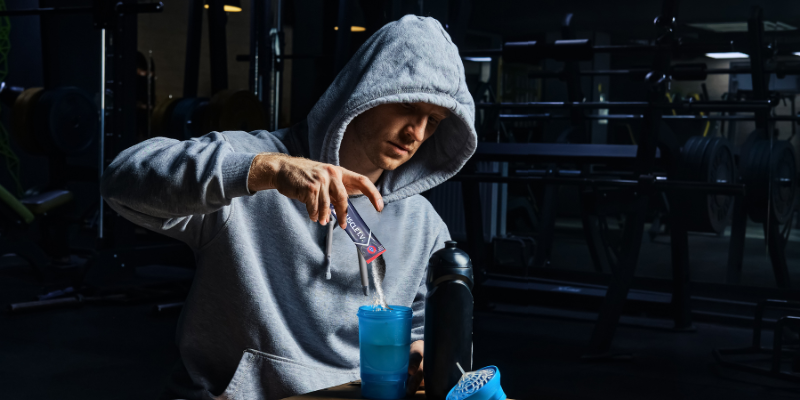 MUSCLE IV homepage image athlete wearing gray hoodie pouring MUSCLE I.V. electrolyte beverage powder into shaker cup.