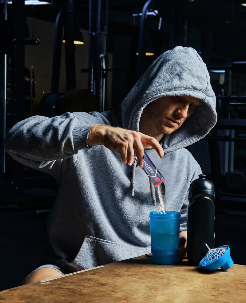 MUSCLE IV homepage image athlete wearing gray hoodie pouring MUSCLE I.V. electrolyte beverage powder into shaker cup.